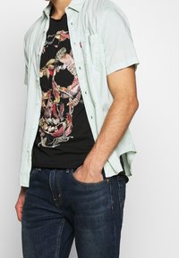 Man wearing light green short-sleeve button-up shirt over black t-shirt with colorful feather skull design and dark blue jeans.