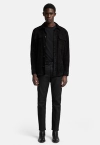 Zwarte suede shirtjack over een zwart t-shirt, gecombineerd met zwarte slim-fit broek en enkellaarsjes. Eenvoudig ontwerp met een gladde textuur.