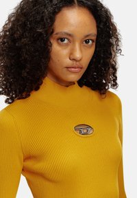 Maglione a collo alto a coste giallo senape con un accento di logo ovale metallico sopra l'area del petto. Texture liscia e design aderente.