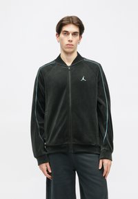 Casaco de veludo preto com fecho zip, apresentando um colarinho alto, detalhes em azul ao longo das mangas e um logotipo da Jordan no peito.