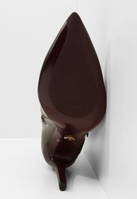 Chaussure à talon aiguille avec un bout pointu, tige en cuir noir et semelle en caoutchouc bordeaux avec des rainures. Détails en métal doré au niveau du talon.