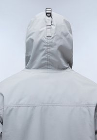 Lys grå hættejakke med et slankt design, glat tekstur og en rem med en metallisk spænde bag på hætten.