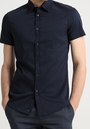 Uomo che indossa una camicia blu navy aderente a maniche corte, abbottonata, infilata in pantaloni scuri con motivo, con le mani nelle tasche.