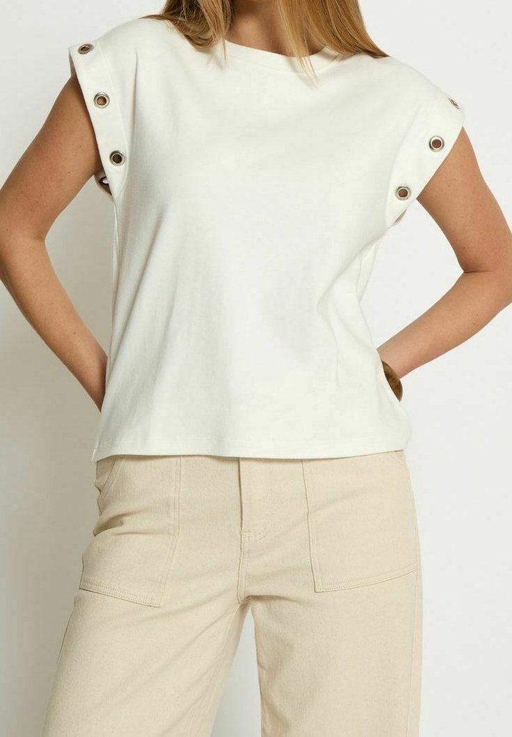 Femme portant un haut blanc sans manches avec des œillets argentés sur les épaules et un pantalon beige avec des poches plaquées à l'avant, debout les mains sur les hanches.