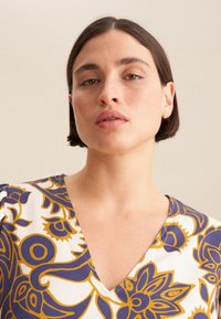 Top a scollo a V in crema con motivo floreale blu e oro. Presenta maniche strutturate e una texture di tessuto liscia, adatta per un abbigliamento casual.