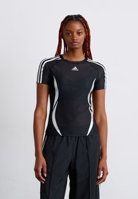 Woman with long red braids standing, wearing black Adidas sports shirt with white stripes and black pants, plain light background.