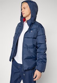 Mørkeblå pufferjakke med hætte, med rød inderside, front zip-lukning og logo på brystet og ærmet.