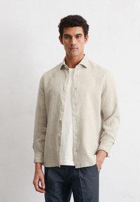 Chemise en lin beige clair avec un devant à boutons, des manches longues et une coupe décontractée, portée par-dessus un t-shirt blanc et assortie à un pantalon sombre.