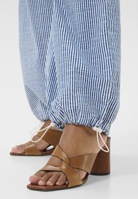 Hnědé kožené sandály na podpatku s křížovými pásky a blokovým podpatkem, doplněné modro-bílými pruhovanými kalhotami s elastickým manžetou.