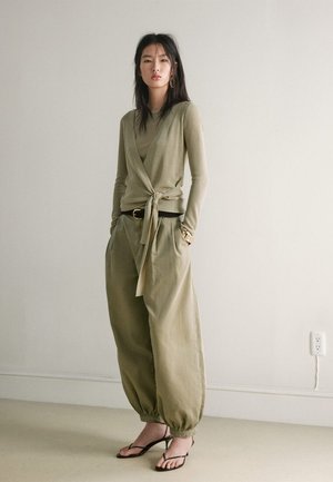 Jeune femme portant un pull cache-cœur olive, pantalon kaki ample resserré aux chevilles, ceinture noire, bracelet doré et sandales noires à talons, debout à l'intérieur.