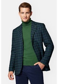 Zielono-granatowa marynarka w kratę z dwoma guzikami, noszona na zielonym golfie. Wyjątkowy detal w postaci agrafki na klapie. Ciemne spodnie dopełniają całość.