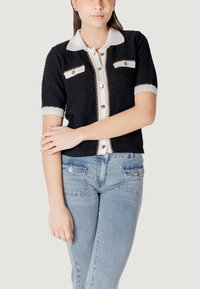 Cardigan corto a maglia nera con colletto bianco e pattina con bottoni. Presenta dettagli con bottoni argentati sulle tasche sul petto. Indossato con jeans azzurri.