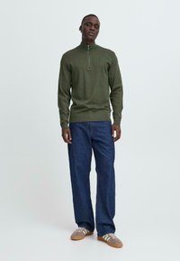 Mørkegrønn strikket genser med halv-zip krage, paret med løstsittende mørkeblå denimbukser og brune joggesko med hvite detaljer.
