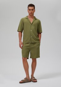 Cămașă scurtă de culoare verde măslin și pantaloni scurți asortați, confecționați dintr-un material texturat, având guler și nasturi. Purtați cu sandale maro.