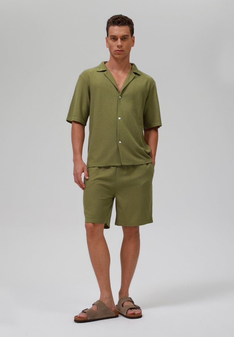 Cămașă scurtă de culoare verde măslin și pantaloni scurți asortați, confecționați dintr-un material texturat, având guler și nasturi. Purtați cu sandale maro.