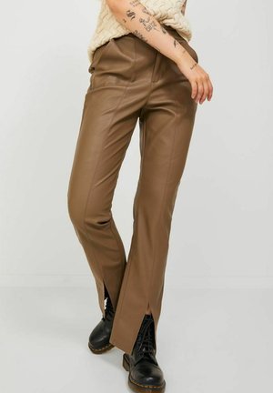 Pantalones de cuero sintético marrón con un diseño ajustado, que presentan aberturas en la parte delantera a la altura de los tobillos. Combinados con botas negras con cordones.