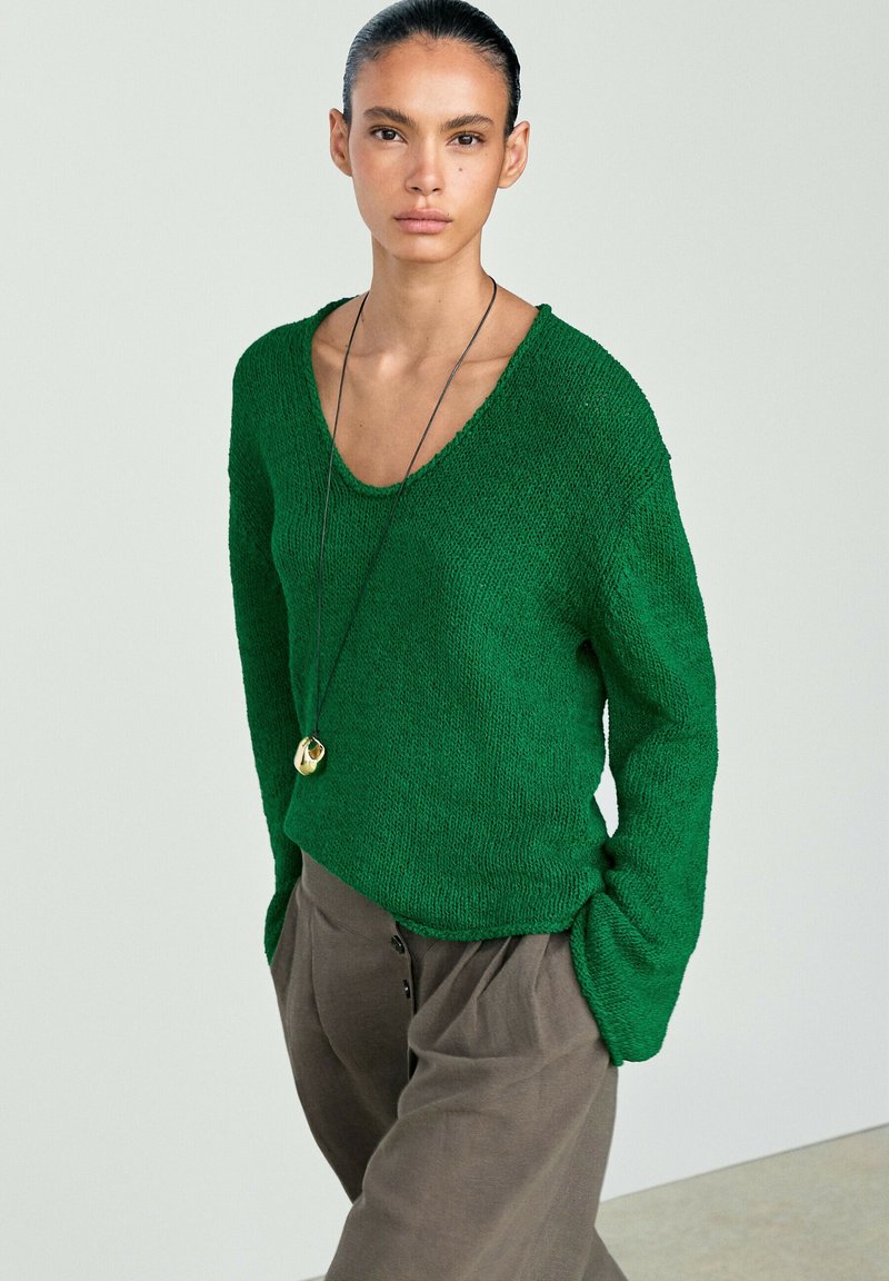 Woman with slicked-back hair wearing a green knit sweater, gray pants, and a long necklace with a gold pendant, standing with hands in pockets.