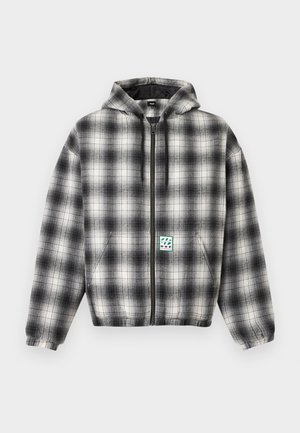 Jeune homme immobile portant un sweat à capuche zippé à carreaux gris et noirs avec un patch logo à l'avant et un pantalon noir avec un design clouté.