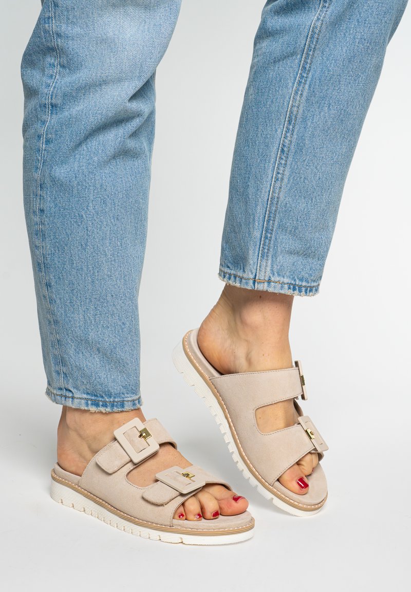 Fødder i lyseblå jeans iført beige slip-on sandaler med to spænder og hvide såler, tæerne malet røde mod en ensfarvet baggrund.