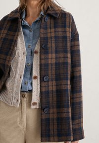 Kobieta ubrana w warstwowy brązowy płaszcz w kratę z wełny, beżowy sweter z dzianiny, niebieską koszulę dżinsową i beżowe spodnie z kordurowej tkaniny.