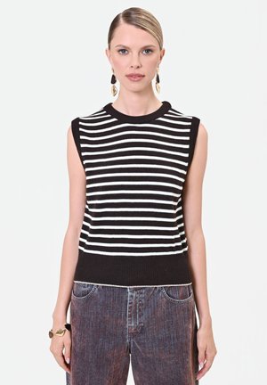 Donna che indossa un top smanicato a righe bianche e nere, jeans scuri lavati, orecchini a goccia dorati e neri e un braccialetto abbinato.