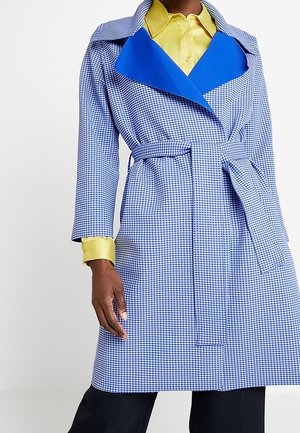 Persona que lleva un abrigo azul a cuadros con cinturón y solapas anchas azules sobre una camisa amarilla abotonada y pantalones negros.