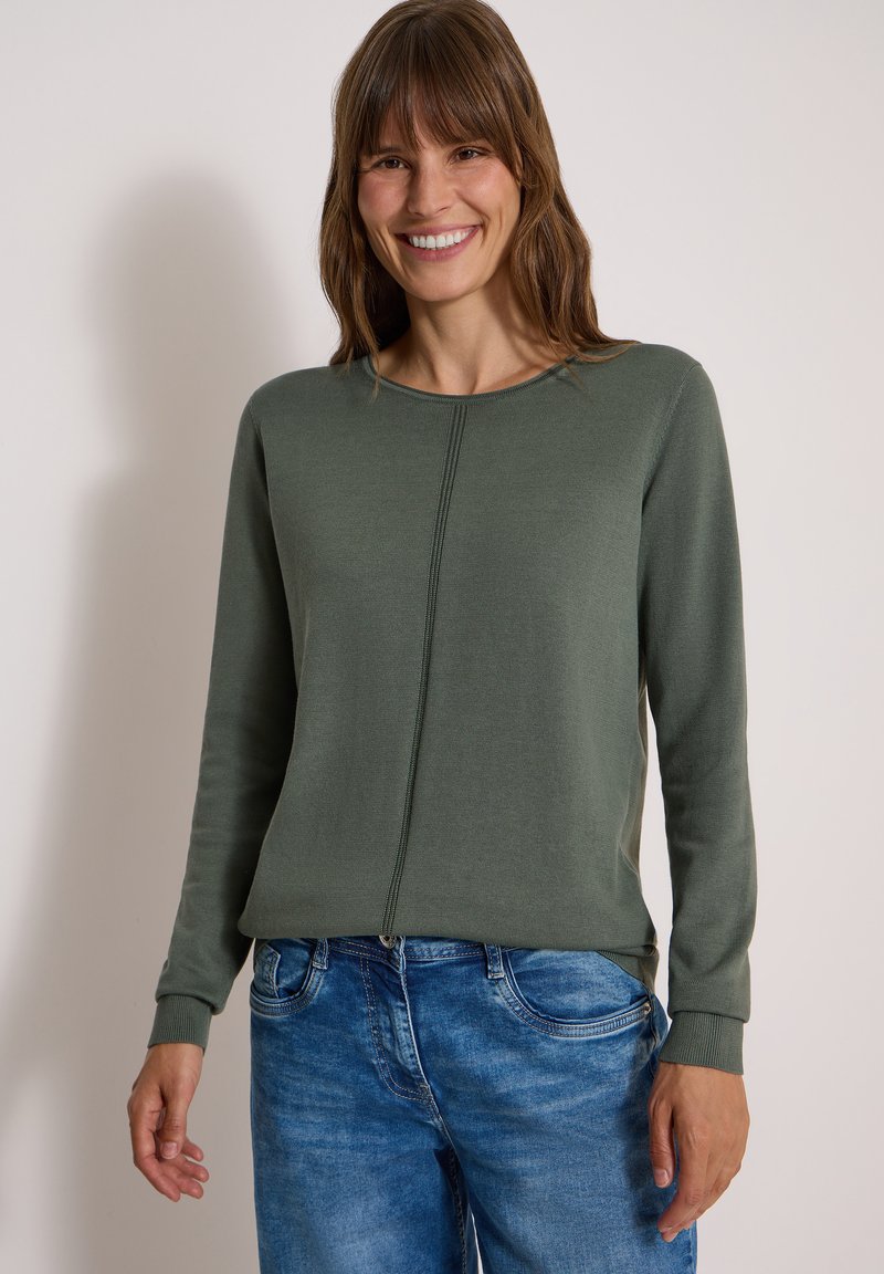 Femme souriante portant un haut vert olive à manches longues et un jean bleu, debout devant un fond clair uni.