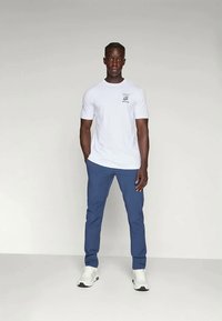 Hvit t-skjorte med korte ermer og liten grafisk design, kombinert med blå bukser. Modellen har på seg hvite joggesko. Enkelt, rene linjer.