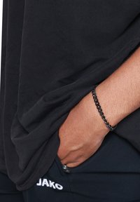 T-shirt nera in cotone con vestibilità ampia, mano infilata in pantaloni neri con marchio "JAKO". Un braccialetto in catena nera è visibile sul polso.