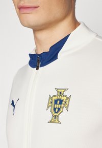 Giacca sportiva bianca con collo blu navy, caratterizzata da un emblema del Portogallo in oro e blu e una zip nera. Tessuto testurizzato con un design sportivo.