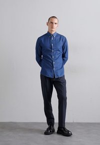 Cămașă albastră cu guler, asortată cu pantaloni închiși și pantofi negri. Materialul pare a fi texturat și neted.
