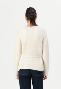 Kvinne med mørkt hår i hestehale iført en kremfarget, fuzzy genser med beltedetalj, kombinert med mørkeblå jeans, stående med ryggen til.