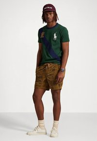 Grön pikétröja med kort ärm, marinblå rand och vitt logo, kombinerad med bruna mönstrade shorts, vita sneakers och en vinröd keps.