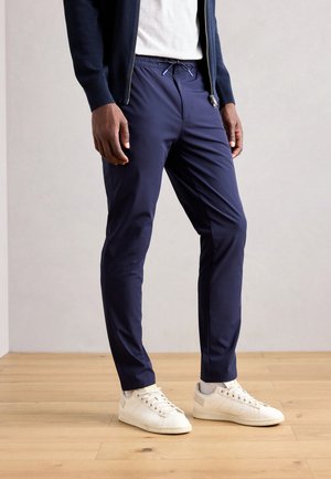 Navy elastic-waist pants with a slim fit, flat front, and subtle sheen. Paired with white lace-up sneakers featuring textured accents.