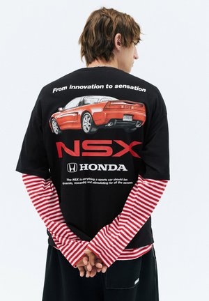 Personne portant un t-shirt noir avec un graphique de voiture rouge, le logo Honda NSX et un texte blanc sur de longues manches rayées rouges et blanches.