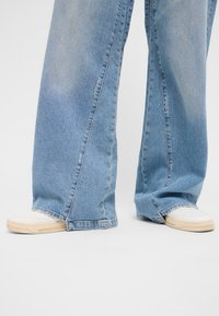 Šviesiai mėlyni plačios kojos džinsai su minkštu paviršiumi ir matomomis siuvimo detalėmis, derinami su baltais sportbačiais, turinčiais perforuotus akcentus.