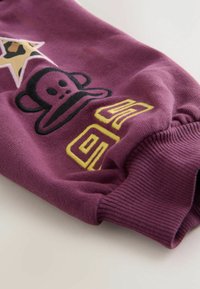 Mâneca unui hanorac violet cu grafică reprezentând o maimuță neagră, accente galbene și manșon cu nervuri. Include un design cu stele și text stilizat. Material moale.