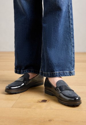 Piedi che indossano mocassini neri lucidi con inserti in camoscio, abbinati a jeans blu a gamba larga, in piedi su un pavimento in legno.