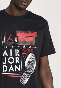 Camiseta de algodón negra con impresiones gráficas en rojo y blanco, incluyendo un diseño de suela de calzado y el texto "AIR JORDAN" y "NIKE AIR."
