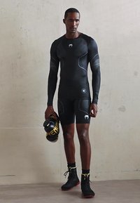 Uomo vestito con abbigliamento sportivo compressivo nero e che tiene guanti da boxe neri e gialli, in piedi di fronte a un muro semplice.