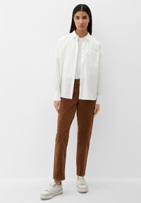 Witte button-up shirt over een witte top, bruine corduroy broek en lichtgekleurde sneakers. Eenvoudig ontwerp, ontspannen pasvorm en geen zichtbare patronen.