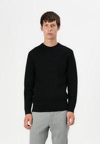 Pull en maille noir avec un col rond et des manches longues, présentant une finition texturée. Associé à un pantalon à rayures claires.