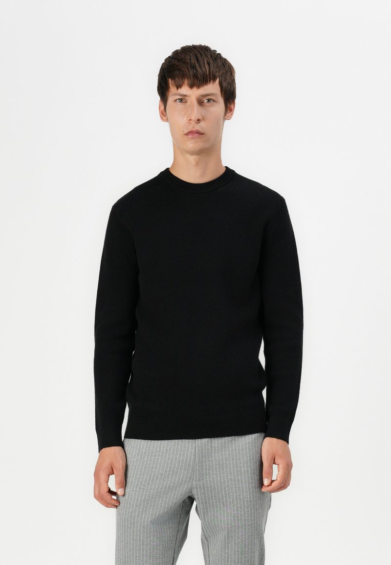 Schwarzer Strickpullover mit rundem Hals und langen Ärmeln, der eine strukturierte Oberfläche aufweist. Kombiniert mit hell gestreiften Hosen.