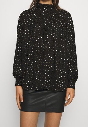 Vrouw die een zwarte blouse met lange mouwen draagt met witte en gouden polkadots, gecombineerd met een zwarte leren rok.