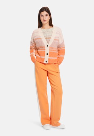 Gestreifter Cardigan in Rosa- und Orangetönen mit einem cremefarbenen Kragen und schwarzen Knöpfen, kombiniert mit orangefarbenen Hosen und weißen Schuhen.