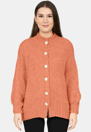 Woman wearing a chunky knitted orange cardigan with large white buttons and black pants, standing against a plain white background.