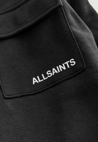 Čierna tkanina s hladkou textúrou; obsahuje vrecko a biely vyšívaný nápis "ALLSAINTS" v pravom dolnom rohu.