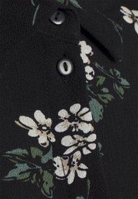 Tissu noir avec imprimé floral blanc et feuilles vertes, avec deux boutons noirs sur un vêtement texturé.