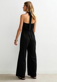 Mono negro con tirantes tipo halter, que presenta un cuerpo ajustado y piernas anchas, con una cintura fruncida y un lazo de satén en el cuello. Textura de tela suave.