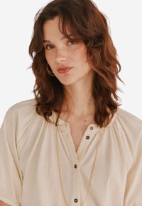 Beige Bluse mit kurzen Ärmeln, geknöpften Ausschnitt, gefältelten Schultern und einem zarten gewellten Saum, akzentuiert durch eine goldene Halskette.